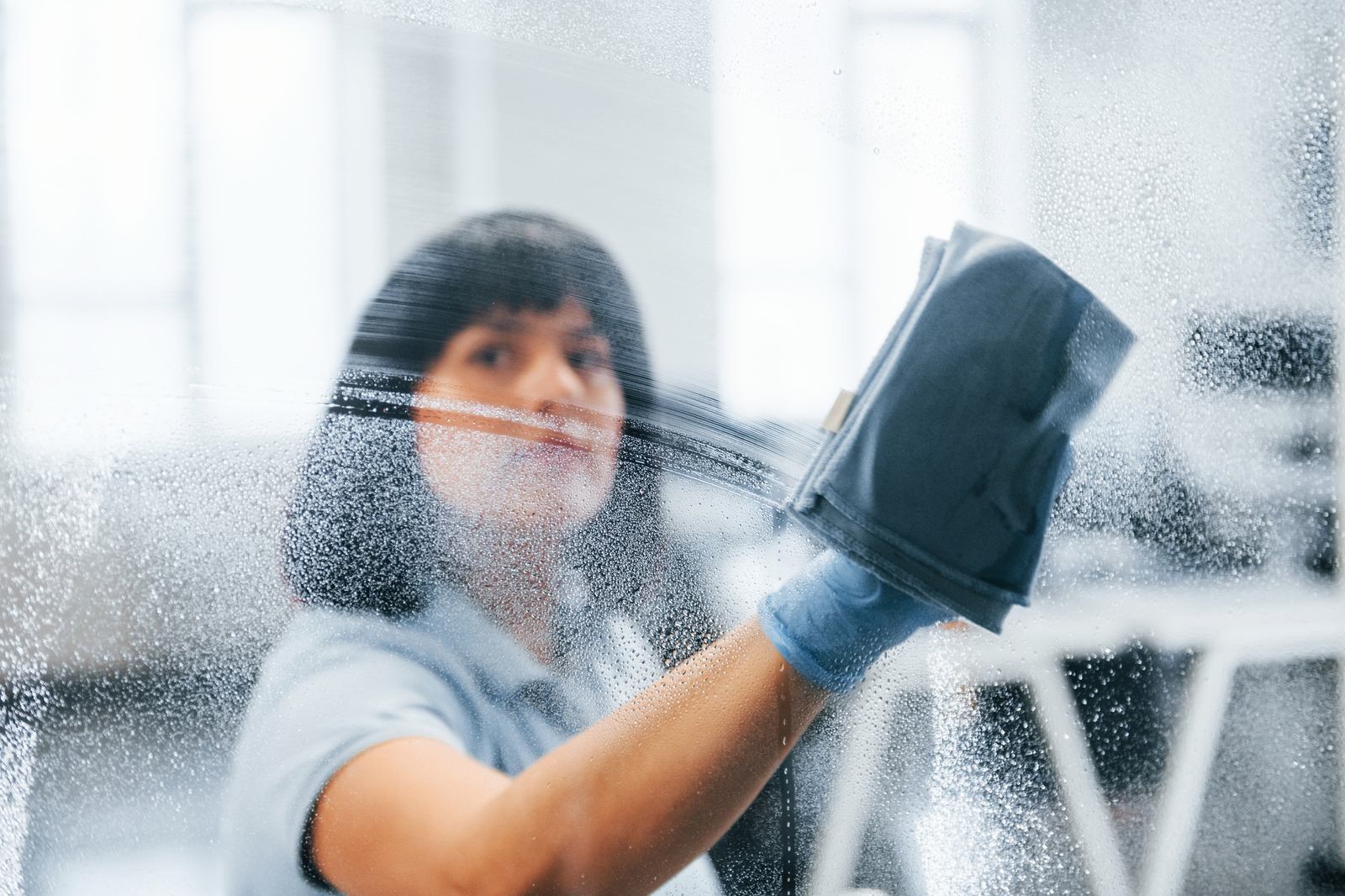 Cleaner in a light blue polo wiping a misted glass pane with a cloth